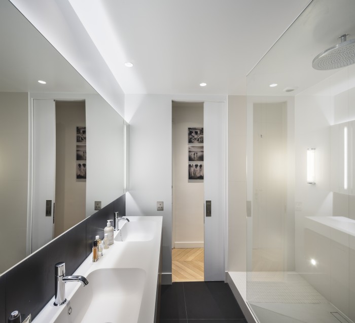 Salle de douche profitant de la lumière naturelle et jouant sur le contraste noir et blanc. Elle est composée d'une porte d'accès à galandage, d'une douche avec double accès et d'une double vasque.